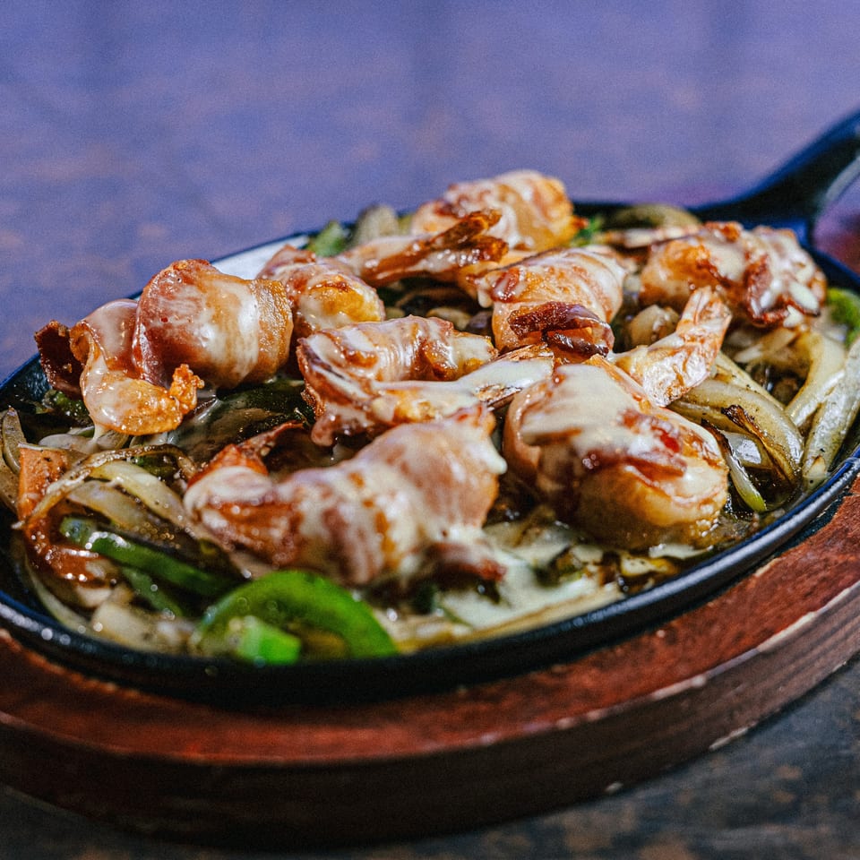 Sizzling fajita skillet with bacon-wrapped shrimp, onions, and green peppers, topped with melted cheese.
