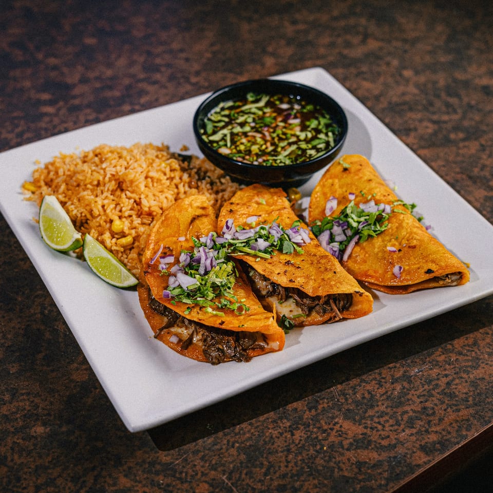 Three birria tacos with rice, lime wedges, and a side of consommé on a white plate.