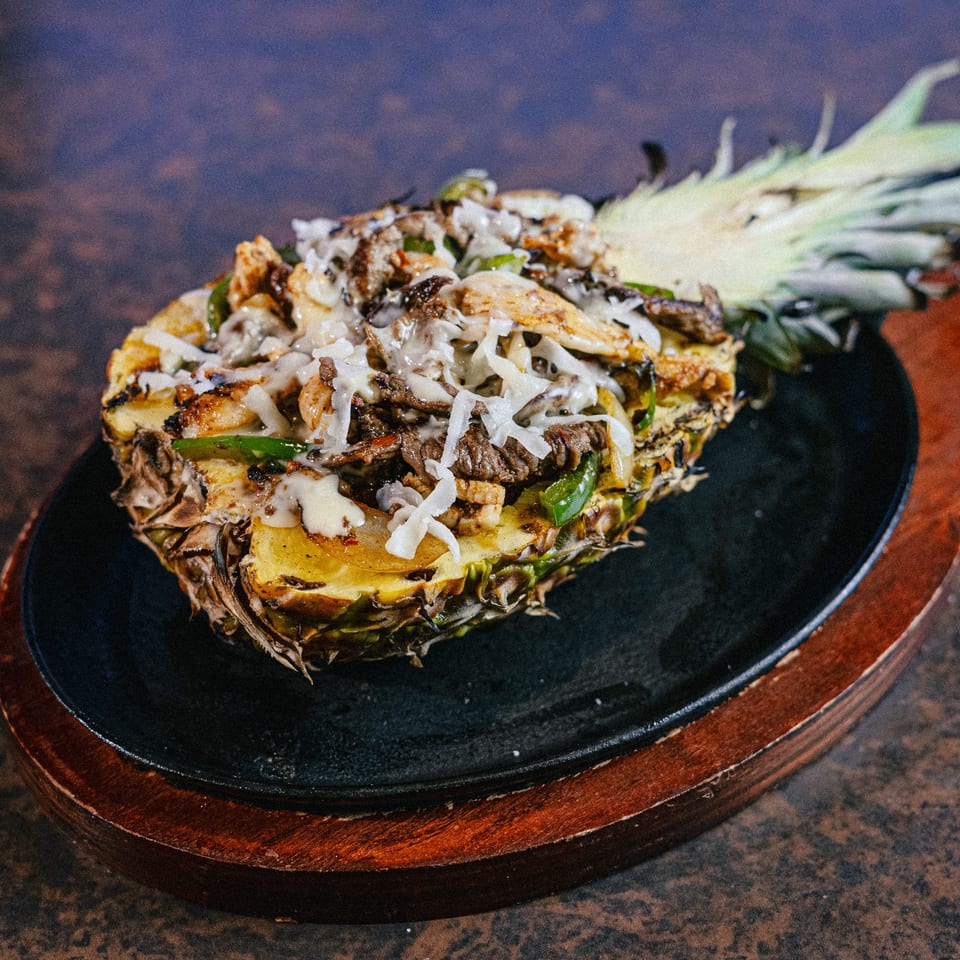Grilled pineapple half filled with steak, chicken, peppers, and melted cheese on a sizzling platter.