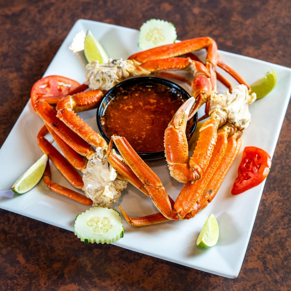 Snow crab legs with a dipping sauce, lime wedges, cucumber, and tomato on a white plate.